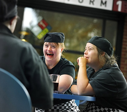  Tre camerieri scherzano seduti intorno ad un tavolino, davanti ad una bibita, in una stazione. Kate, una di loro, ha la sindrome di Down. © Richard Bailey